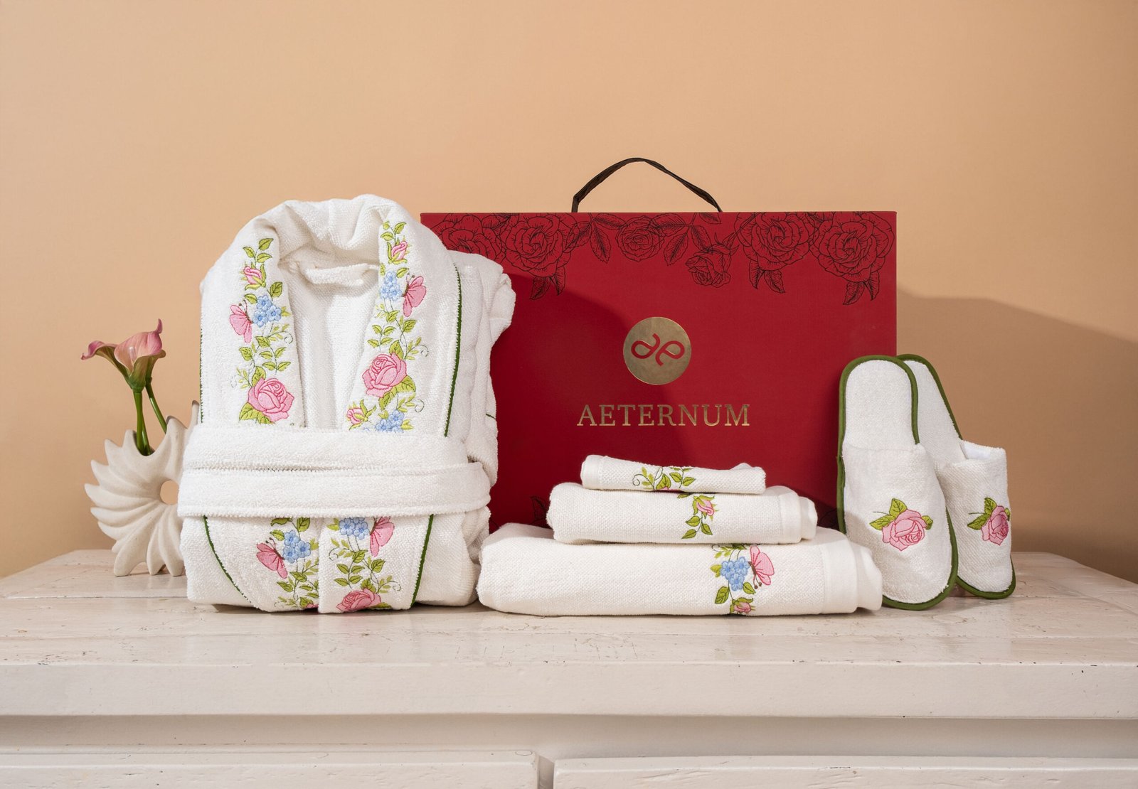 White embroidered bathrobe, folded towels and slippers arranged beside an Aeternum gift box
